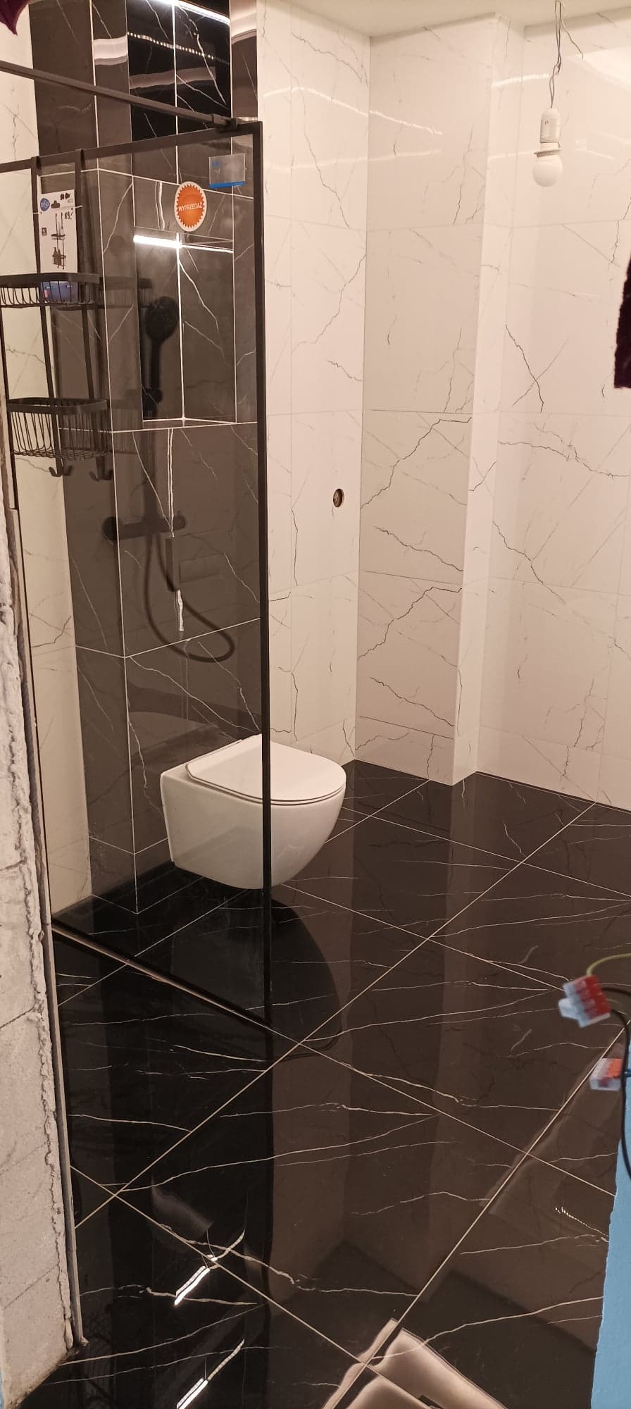 Wykończona łazienka z czarno-białymi płytkami imitującymi marmur, podwieszana toaleta i kabina prysznicowa z czarnymi profilami.