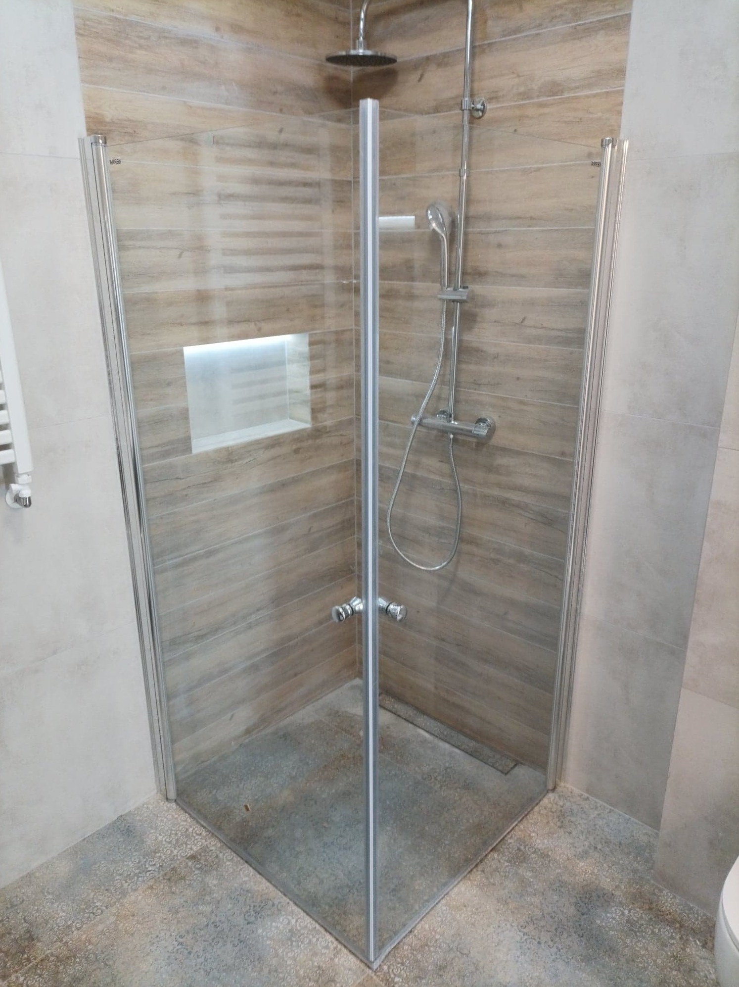 Kabina prysznicowa z chromowanymi elementami i szklanymi drzwiami, wykończona płytkami imitującymi drewno, z wnęką oświetleniową w ścianie.