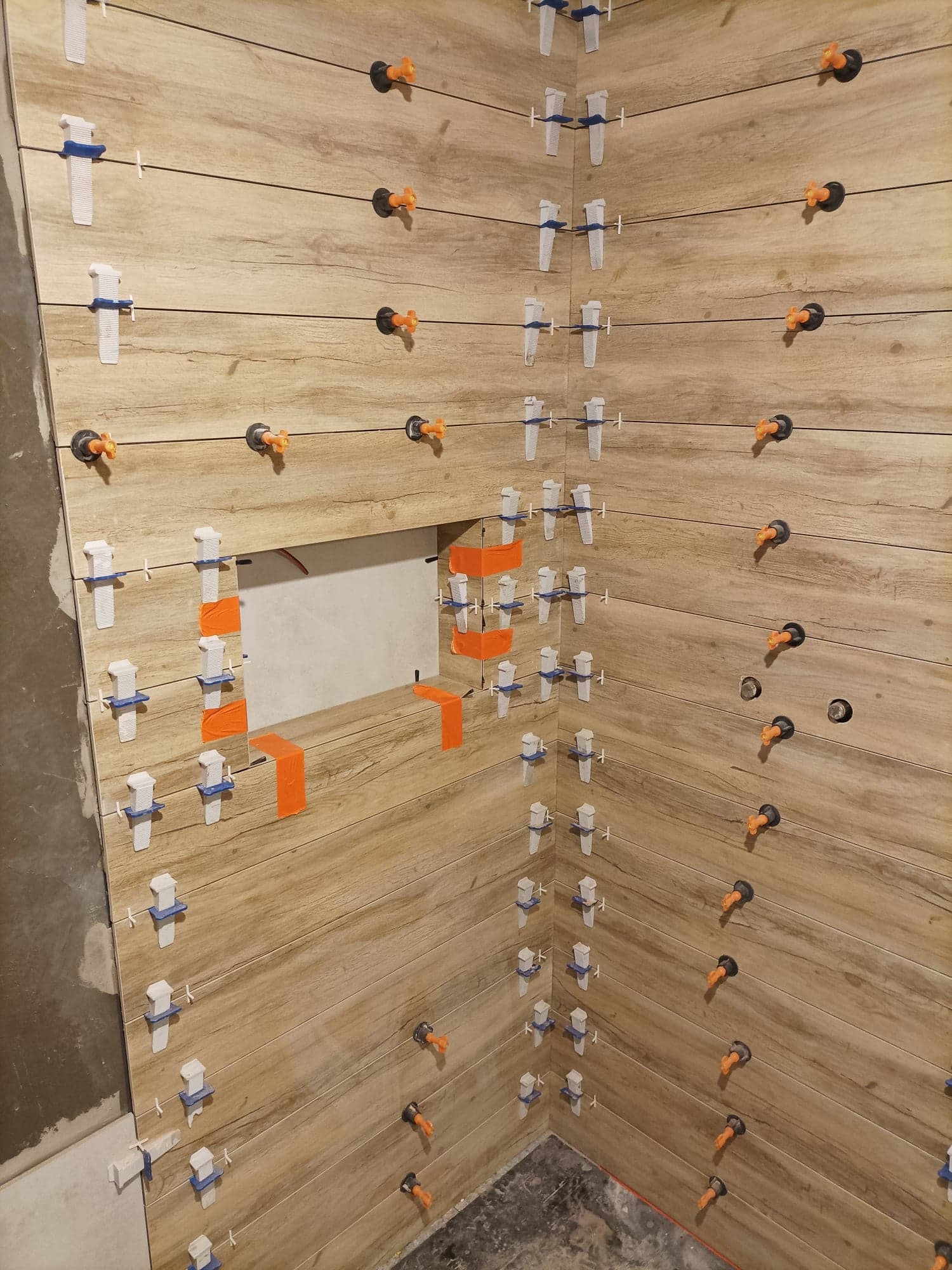 Układanie płytek imitujących drewno w rogu łazienki, widoczne klipsy i kliny poziomujące oraz pomarańczowa taśma zabezpieczająca otwór rewizyjny.