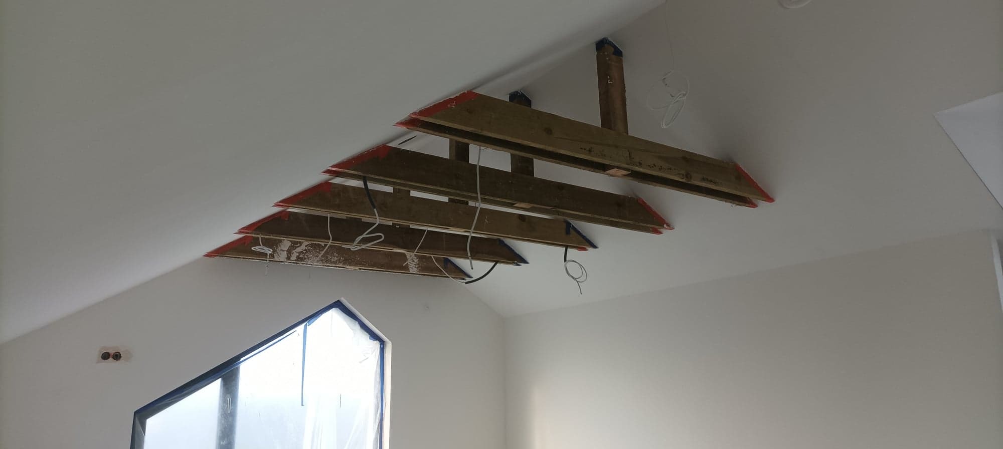 Montaż drewnianej konstrukcji pod sufitem w nowym domu, widoczne kable i elementy instalacji elektrycznej, okno zabezpieczone folią.