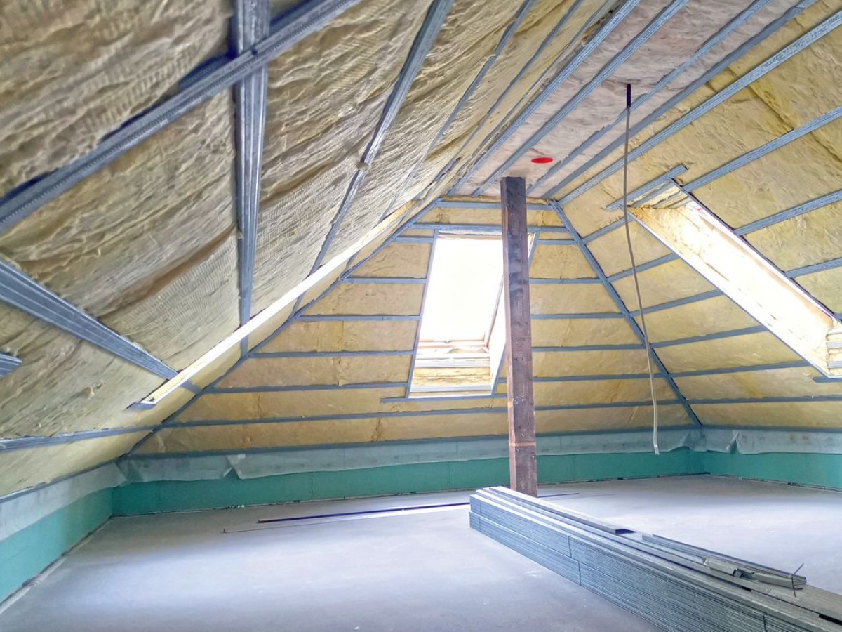 Ocieplenie poddasza z oknem dachowym w Skierniewicach, widoczne metalowe profile i wełna mineralna, na podłodze stos metalowych elementów konstrukcyjnych.