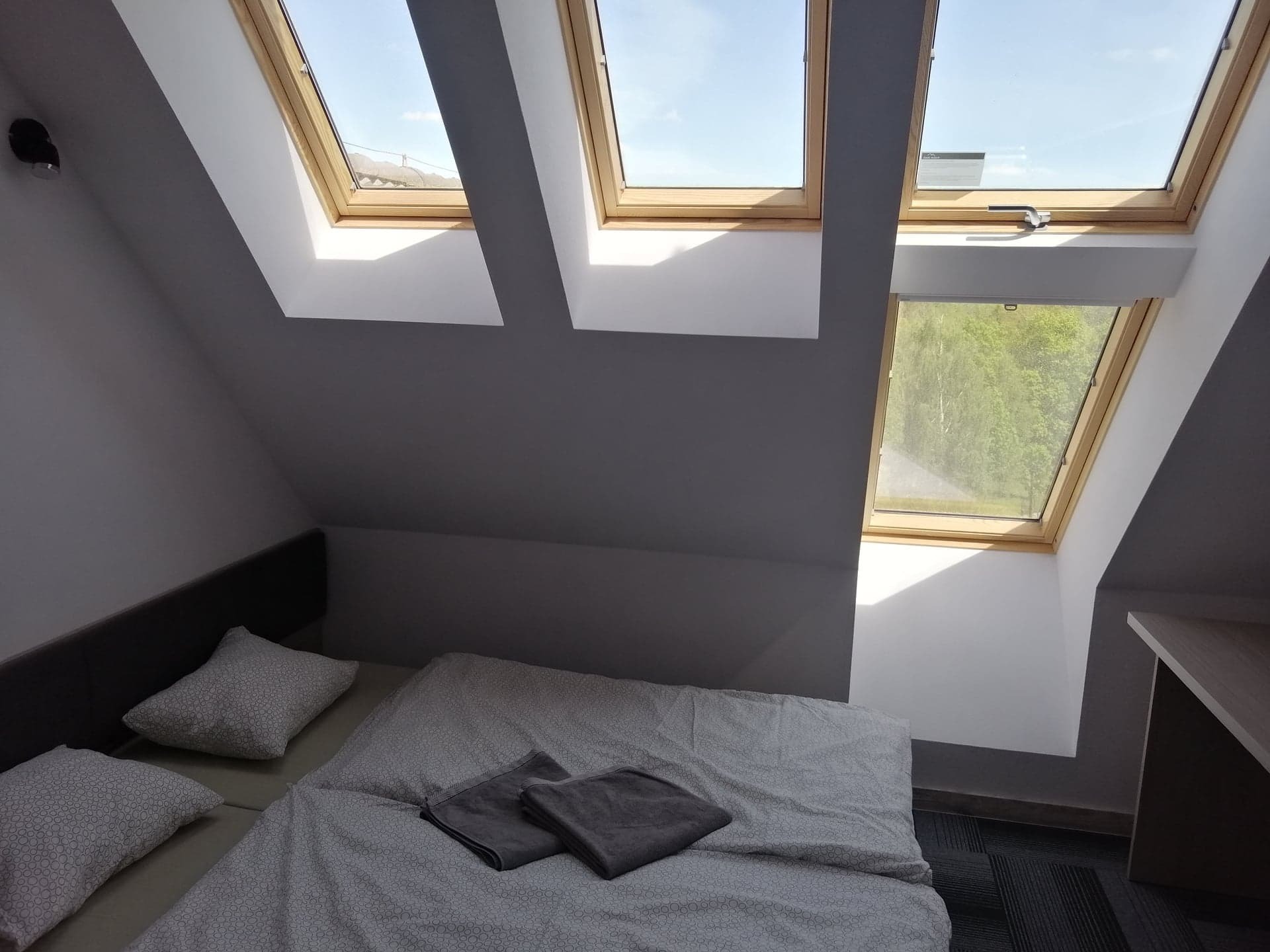 Pokój na poddaszu z łóżkiem i trzema oknami dachowymi z widokiem na błękitne niebo i zieleń, ściany pomalowane na szaro.