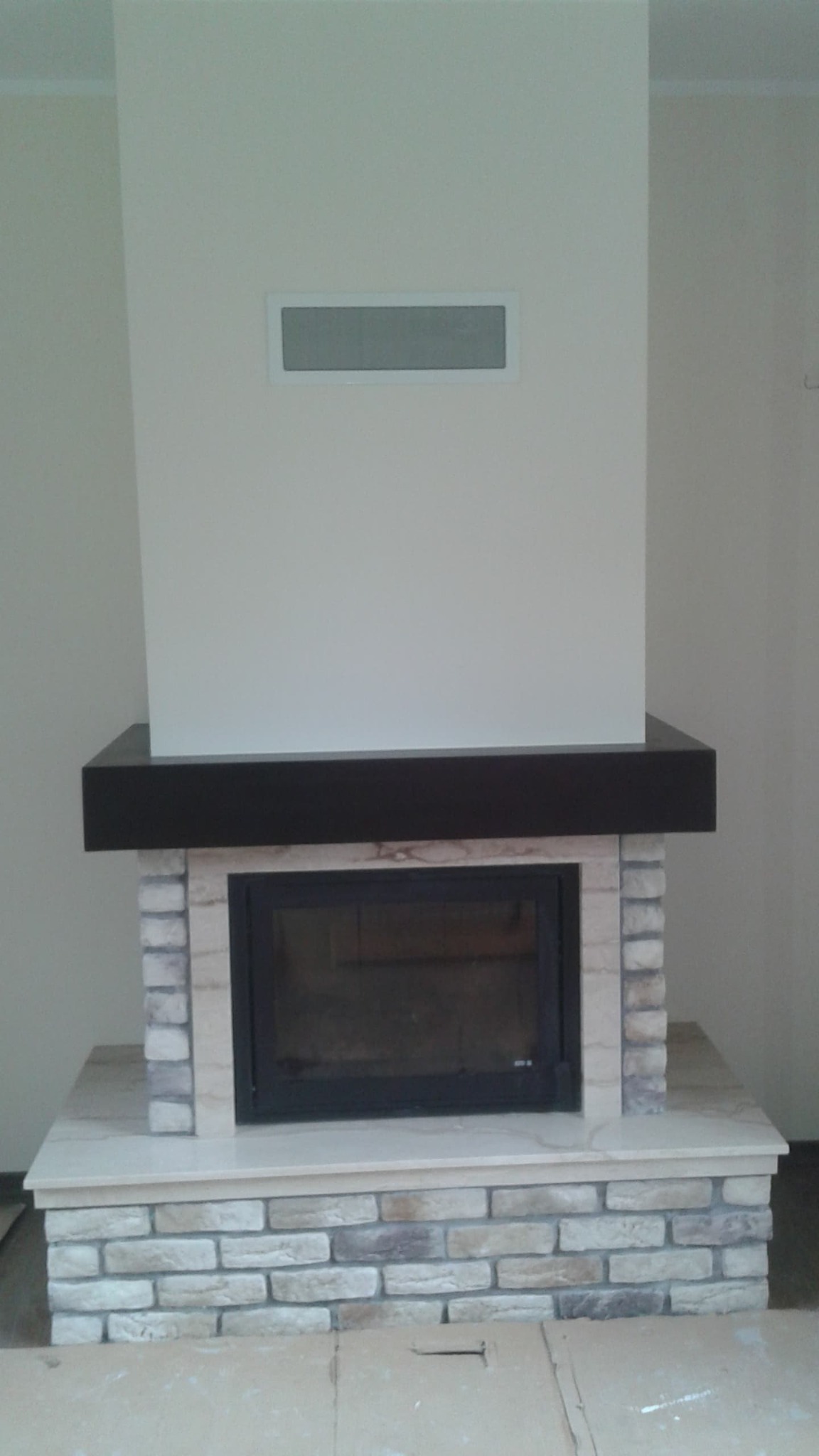 Nowoczesny kominek obudowany cegłą dekoracyjną w odcieniach szarości i beżu, z ciemnym drewnianym blatem, umieszczony na tle jasnej ściany z kratką wentylacyjną.