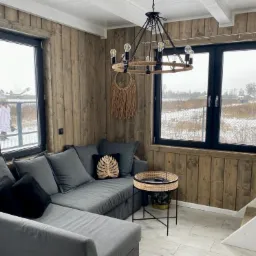 Wnętrze domu z drewnianą boazerią, szary narożnik z poduszkami, okrągły stolik z rattanowym wykończeniem, widok za oknem na zimowy krajobraz i bawiące się dzieci.