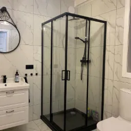 Nowoczesna kabina prysznicowa z czarnymi profilami w łazience wykończonej białymi płytkami imitującymi marmur, widoczna umywalka z szafką i okrągłe lustro.