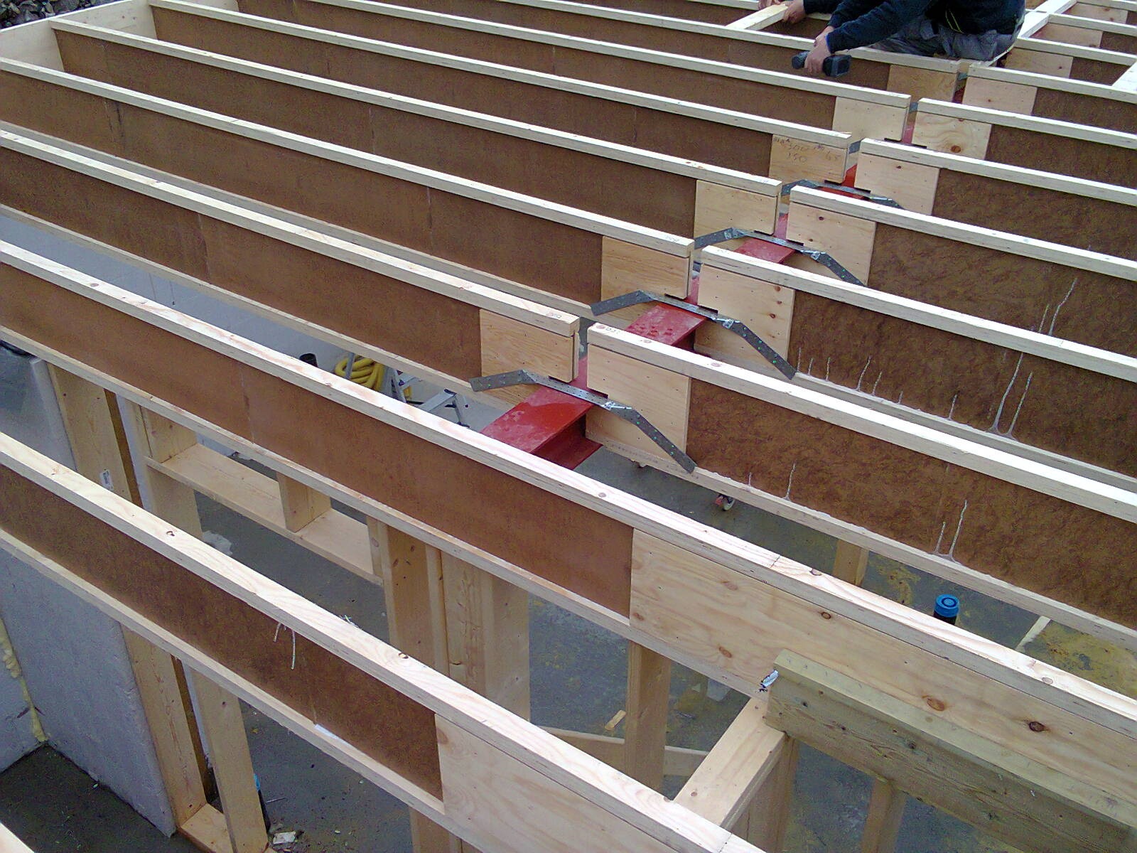 Konstrukcja szkieletowa podłogi domu w trakcie montażu, widoczne drewniane belki, płyty OSB oraz metalowe łączniki, częściowo ocieplona wełną mineralną.