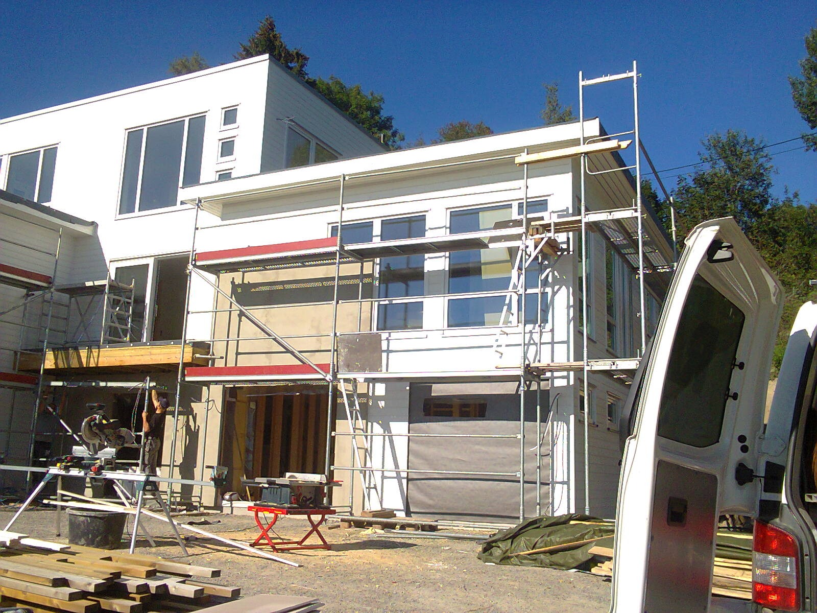 Budowa nowoczesnego, dwukondygnacyjnego domu z białymi ścianami, widoczne rusztowania, otwarte drzwi garażowe zabezpieczone szarą plandeką, narzędzia budowlane i otwarty samochód dostawczy z boku.