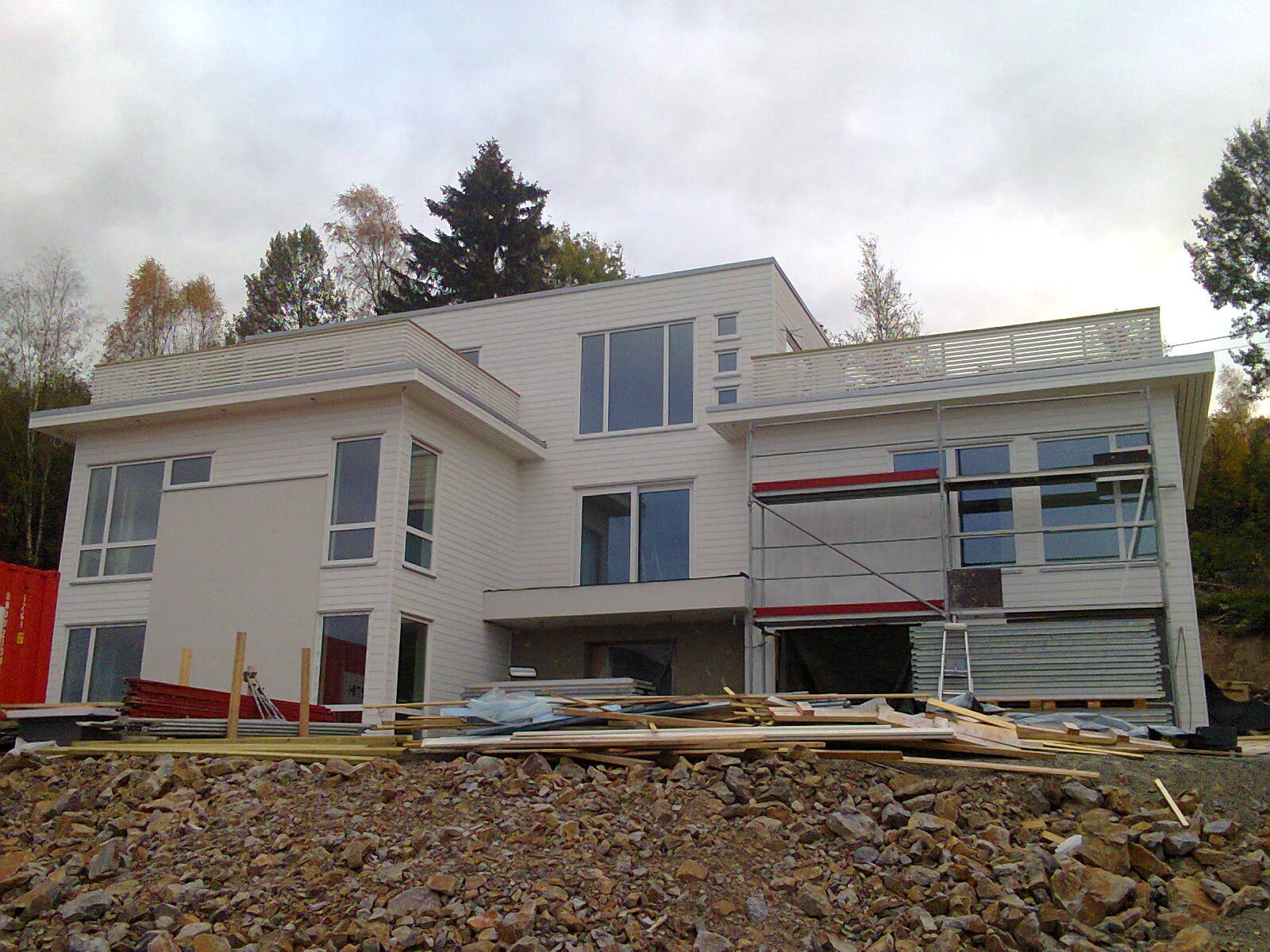 Nowoczesny, biały dom w trakcie budowy, widoczne rusztowania i materiały budowlane na pierwszym planie, zieleń drzew w tle.