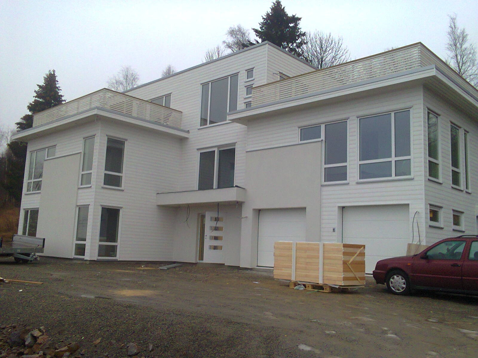 Nowoczesny, dwukondygnacyjny dom z białym sidingiem i płaskim dachem, z balkonami na dachu, widoczny z zewnątrz, z zaparkowanym samochodem i paletą drewna przed garażem, w pochmurny dzień.