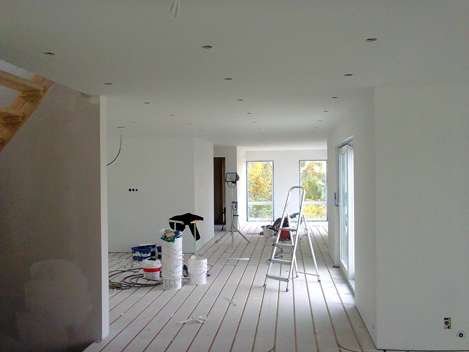 Wnętrze domu w trakcie wykańczania: białe ściany, podłoga z ogrzewaniem podłogowym, drabina, puszki z farbą i okna z widokiem na jesienne drzewa.