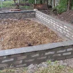 Wykopany fundament z częściowo wymurowanymi ścianami z szarej cegły, wypełniony ziemią, widoczne zabezpieczenie hydroizolacyjne, teren budowy.