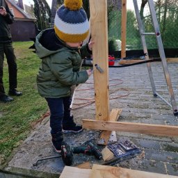 GEPOLDE Sp. z o.o. - Dziecko w czapce z pomponem przykręca śrubę w drewnianej konstrukcji na zewnątrz, w tle drabina i budynki mieszkalne.