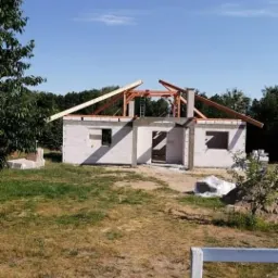 Wznoszenie ścian parterowego domu z betonu komórkowego; widoczne drewniane elementy konstrukcji dachu oraz drabina oparta o ścianę szczytową.