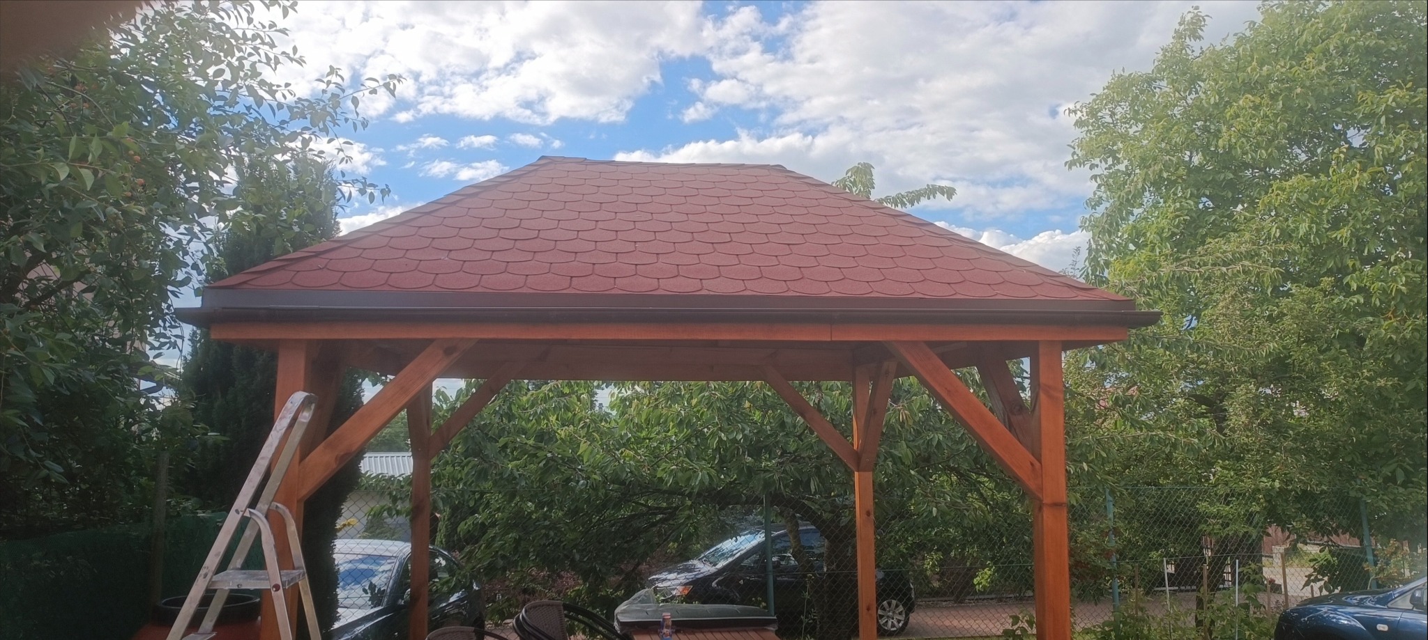 Drewniana altana ogrodowa z czerwonym dachem bitumicznym, widoczna drabina oparta o konstrukcję, otoczona zielenią drzew i krzewów.