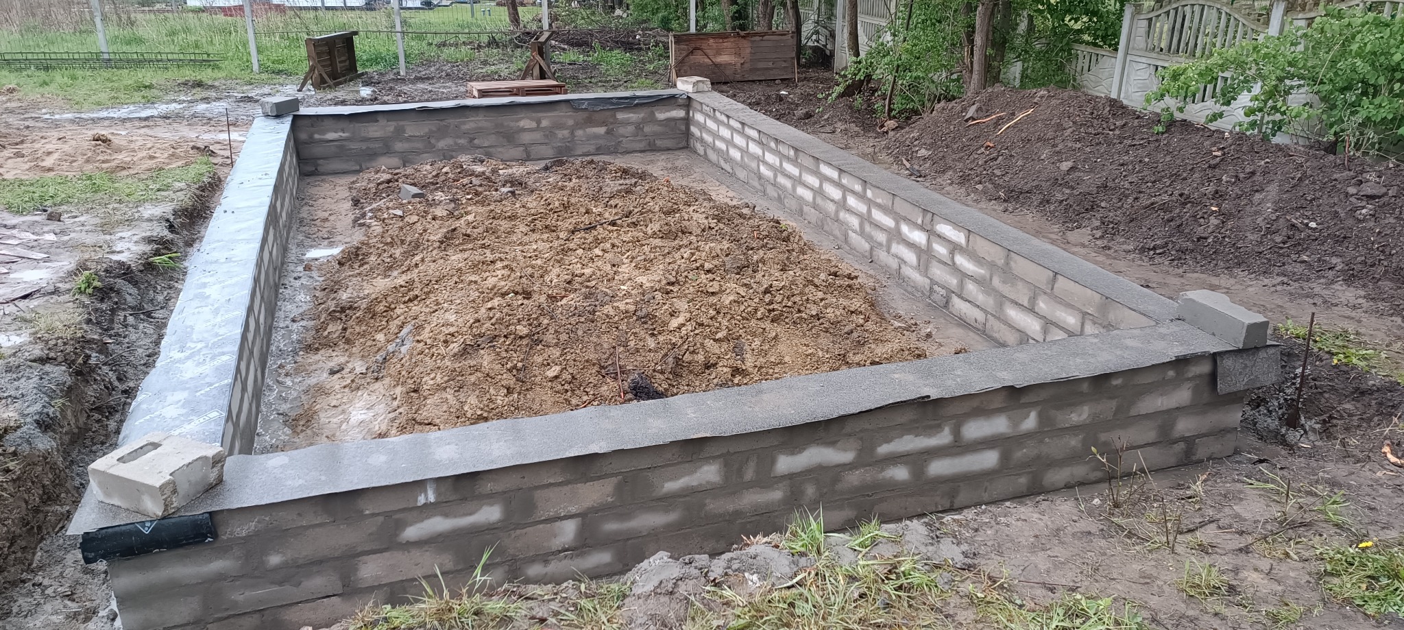 Wykopany fundament z częściowo wymurowanymi ścianami z szarej cegły, wypełniony ziemią, widoczne zabezpieczenie hydroizolacyjne, teren budowy.