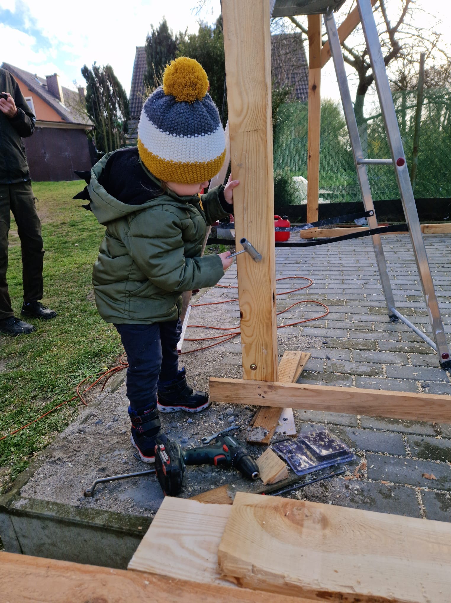 Dziecko w czapce z pomponem przykręca śrubę w drewnianej konstrukcji na zewnątrz, w tle drabina i budynki mieszkalne.