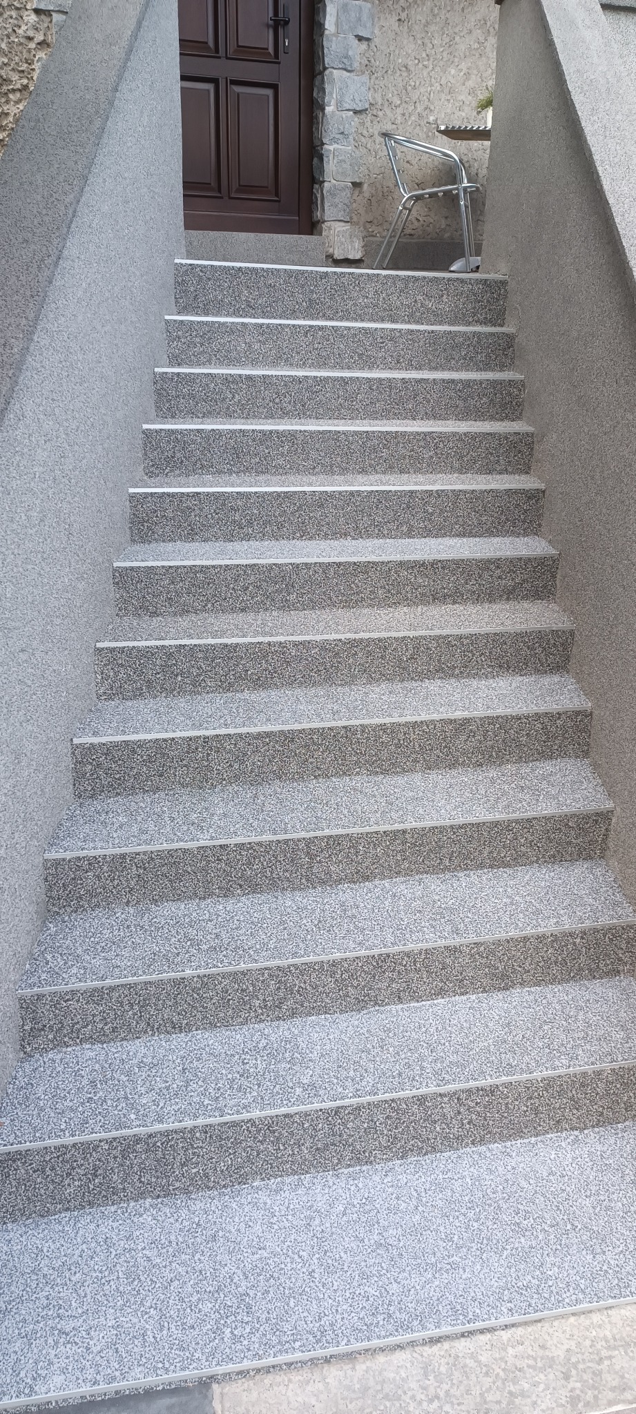 Zewnętrzne schody wejściowe do domu wykończone szarą, kamyczkową fakturą, z widocznymi aluminiowymi listwami zabezpieczającymi krawędzie stopni.