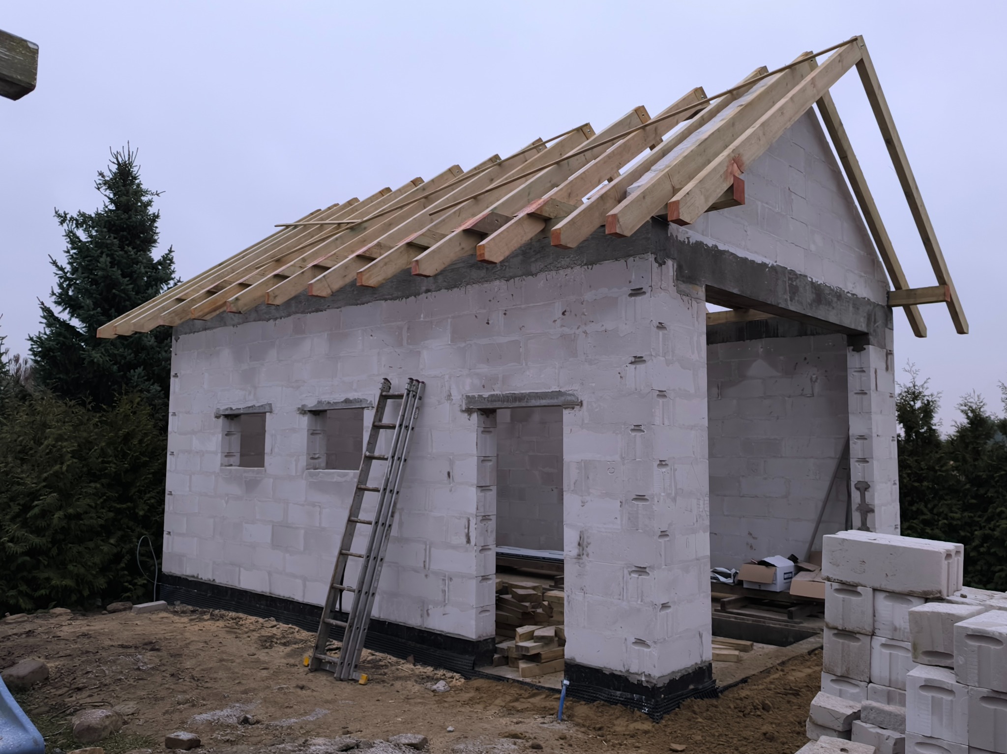 Budowa domu z betonu komórkowego w stanie surowym otwartym, widoczne ściany, otwory okienne i drzwiowe, drewniana konstrukcja dachu oraz drabina oparta o ścianę.
