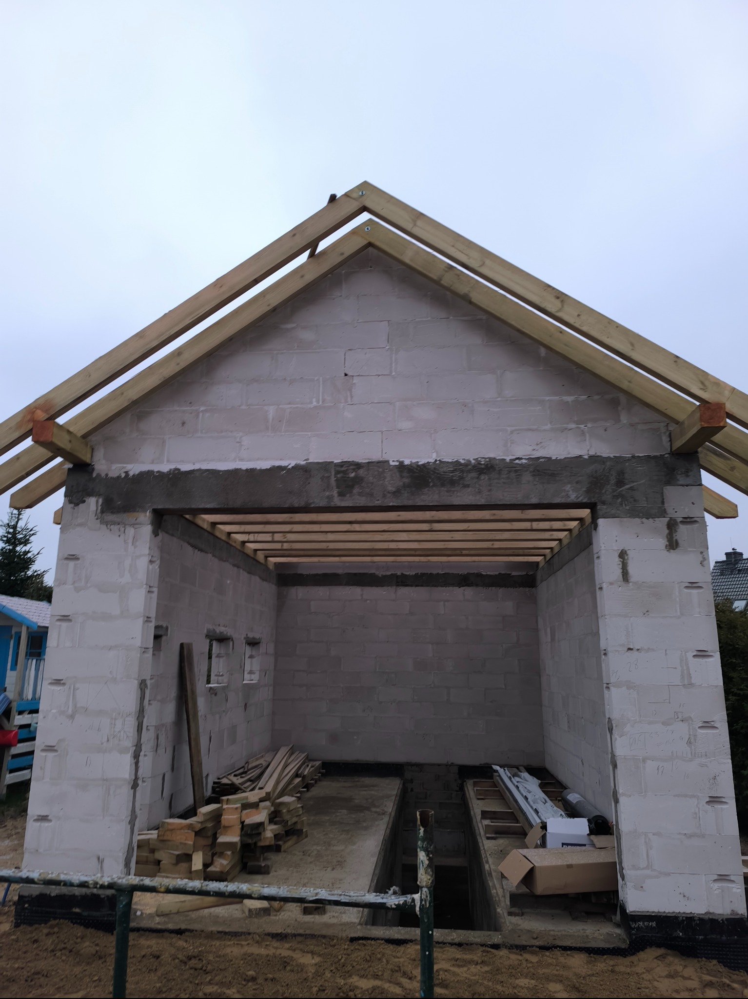 Surowa konstrukcja garażu z betonu komórkowego, z widocznymi belkami dachowymi i otworem na bramę, wewnątrz widoczny kanał samochodowy.