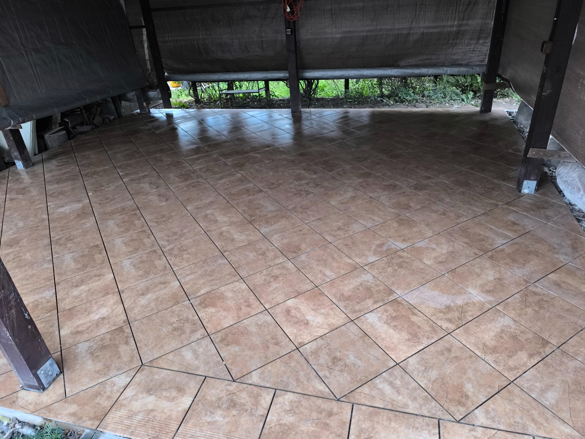 Ułożona, brązowa terakota na podłodze zadaszonego tarasu. Widoczne drewniane słupy konstrukcyjne i zieleń w tle. Ułożenie płytek w karo.