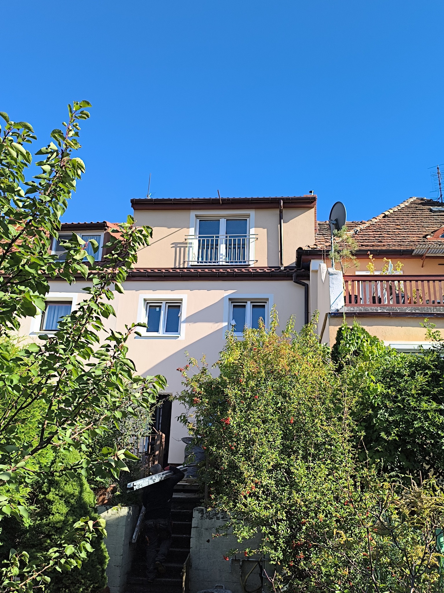 Dwupiętrowy dom z balkonem, widziany z dołu przez bujną zieleń ogrodu. Jasna elewacja, białe okna i brązowy dach kontrastują z intensywnym błękitem nieba.