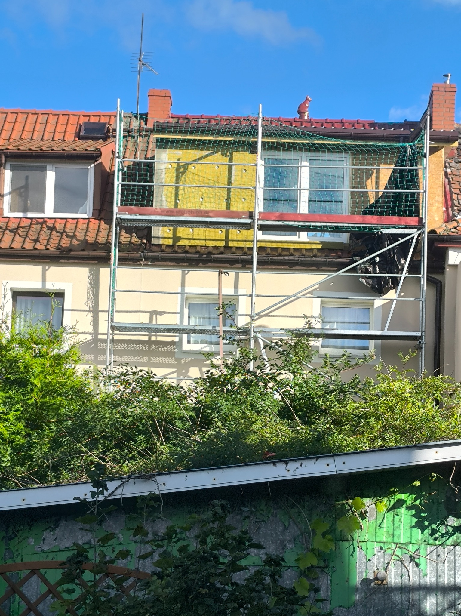 Ocieplanie ściany budynku z rusztowaniem, widoczna żółta izolacja. Na dachu czerwona dachówka i ozdobna figurka kota. Ujęcie z dołu, zza zieleni.