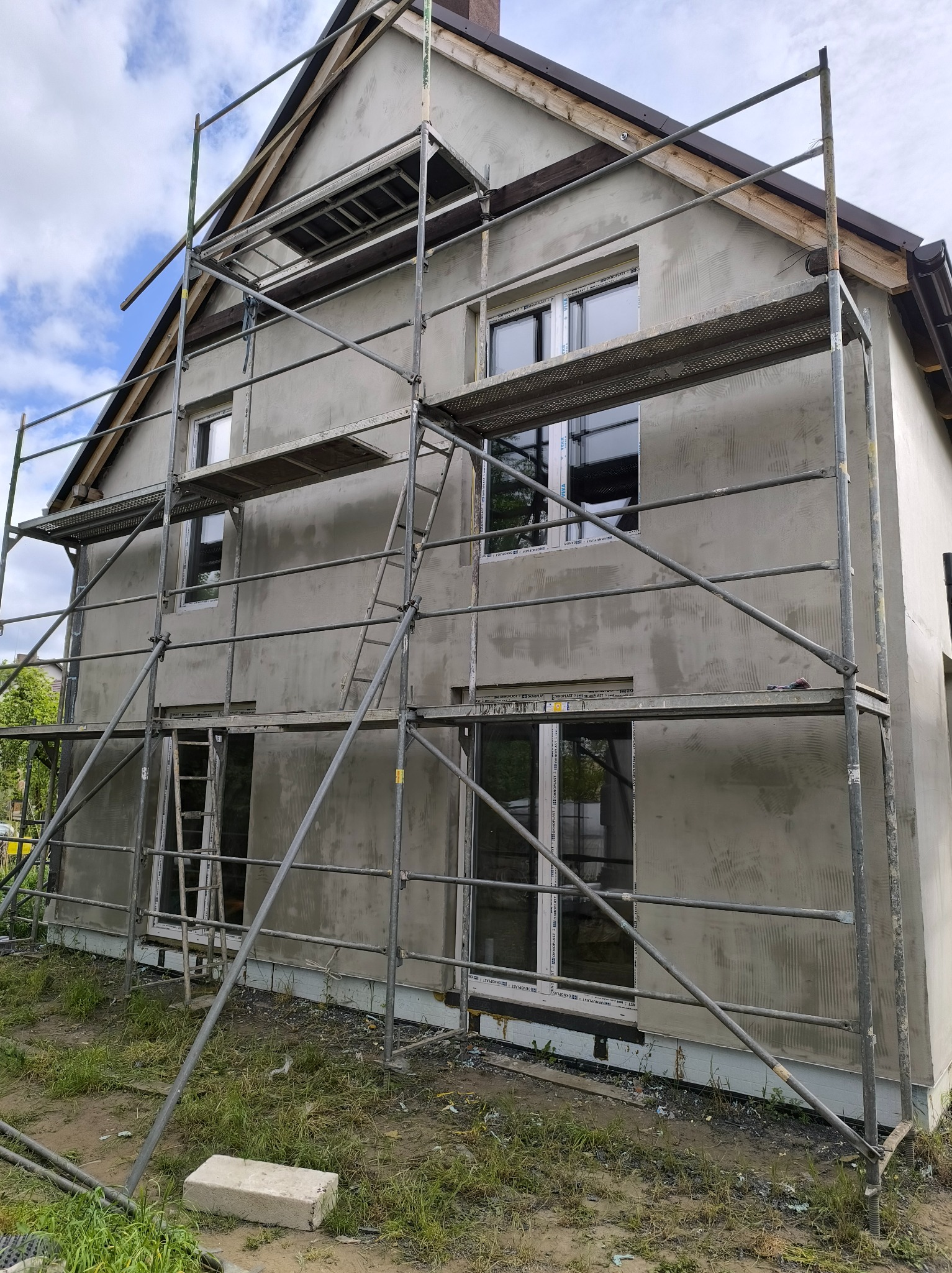 Dwukondygnacyjny dom w trakcie prac elewacyjnych, z widocznymi rusztowaniami i nałożoną warstwą szarego tynku. Widoczne okna z białymi ramami i fragment dachu.