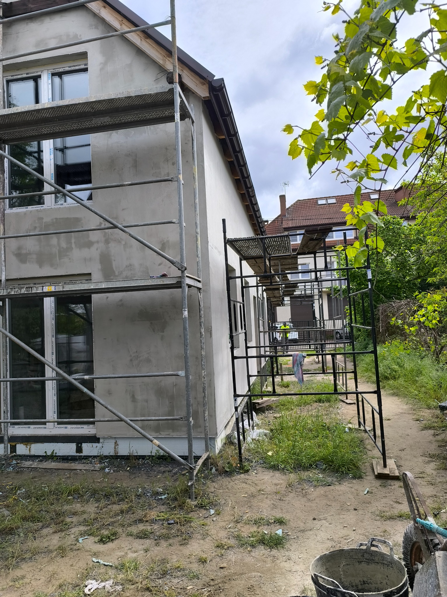 Dwukondygnacyjny dom w trakcie ocieplania elewacji, z widocznymi rusztowaniami i nałożoną warstwą szarego tynku. W tle zieleń i sąsiednie budynki.
