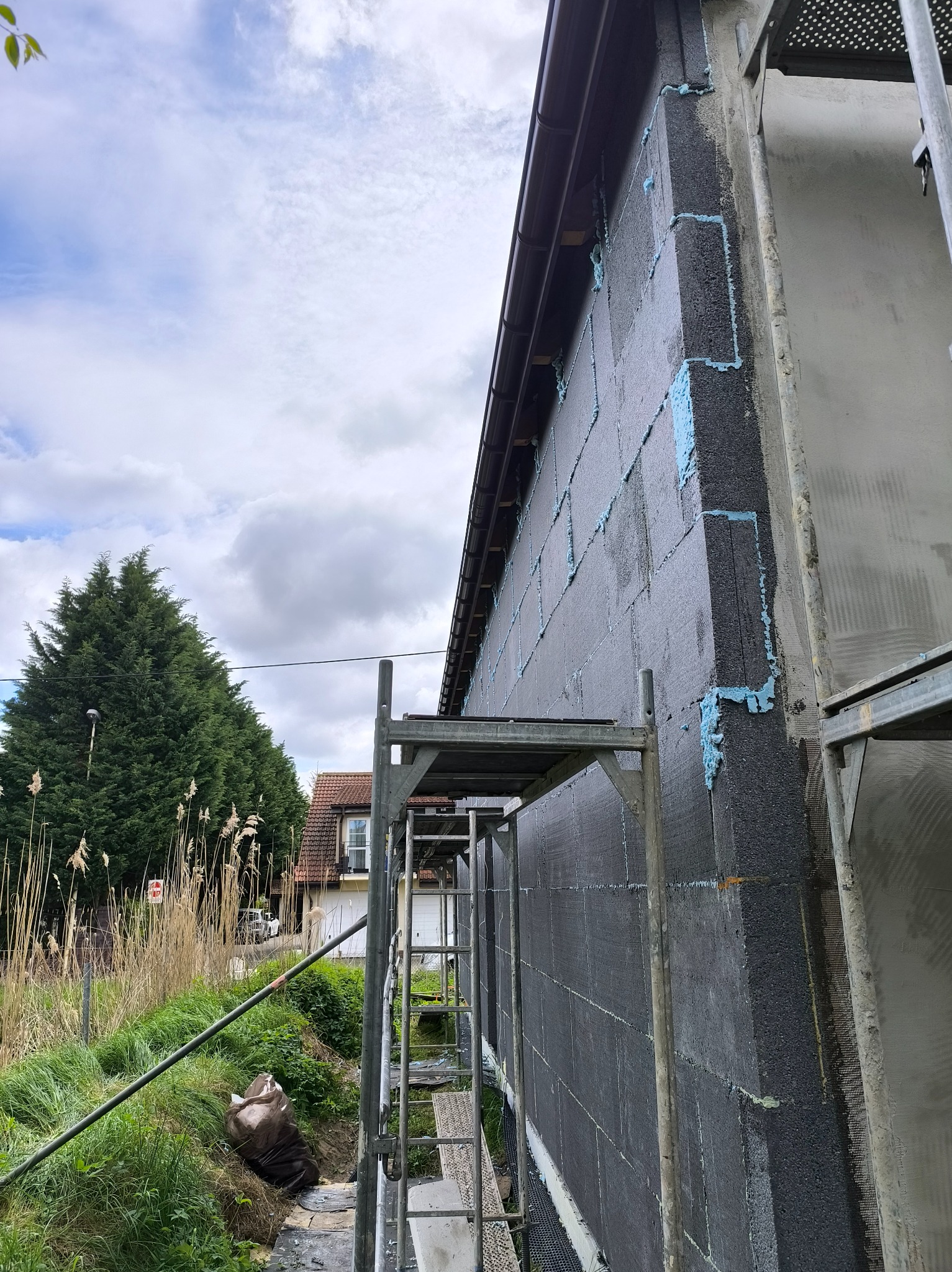 Ściana budynku w trakcie ocieplania, widoczne bloczki z szarego styropianu połączone pianką montażową. Rusztowanie przy ścianie. Ujęcie z dołu, widok na niebo.