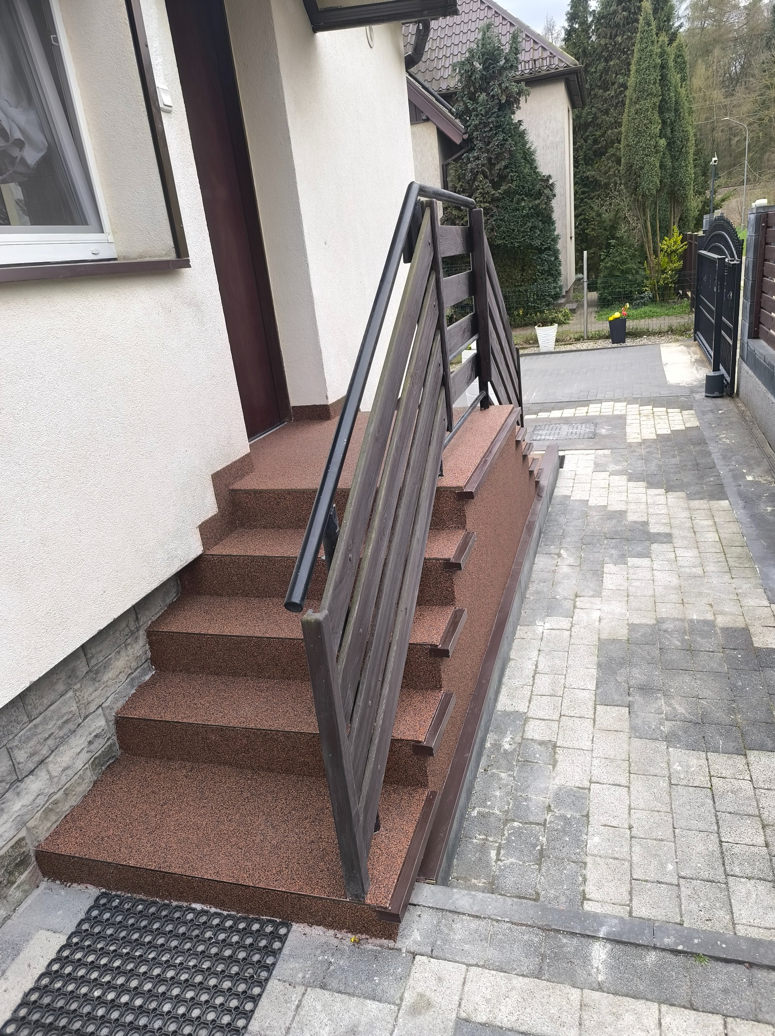 Zewnętrzne schody wejściowe do domu wykończone brązowym, żywicznym materiałem. Widoczne barierki i fragment kostki brukowej. Ujęcie z boku, podkreślające detale wykończenia.