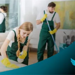 Trzy osoby w zielonych kombinezonach i ż&oacute;łtych rękawicach sprzątają wnętrze: kobieta przeciera blat, mężczyzna myje podłogę, a kolejna kobieta czyści okno; w prawym dolnym rogu logo firmy...