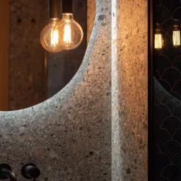 Nowoczesna łazienka z umywalką, baterią w kolorze czarnym, ścianą z terazzo i okrągłym lustrem podświetlonym żarówkami Edisona.