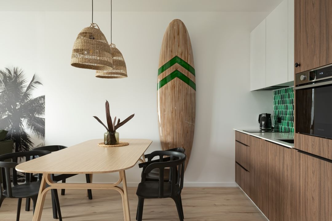 Jasna jadalnia z drewnianym stołem i czarnymi krzesłami, nad stołem wiszą trzy plecione lampy, w tle deska surfingowa z zielonymi paskami oparta o ścianę, fragment kuchni z brązowymi szafkami...