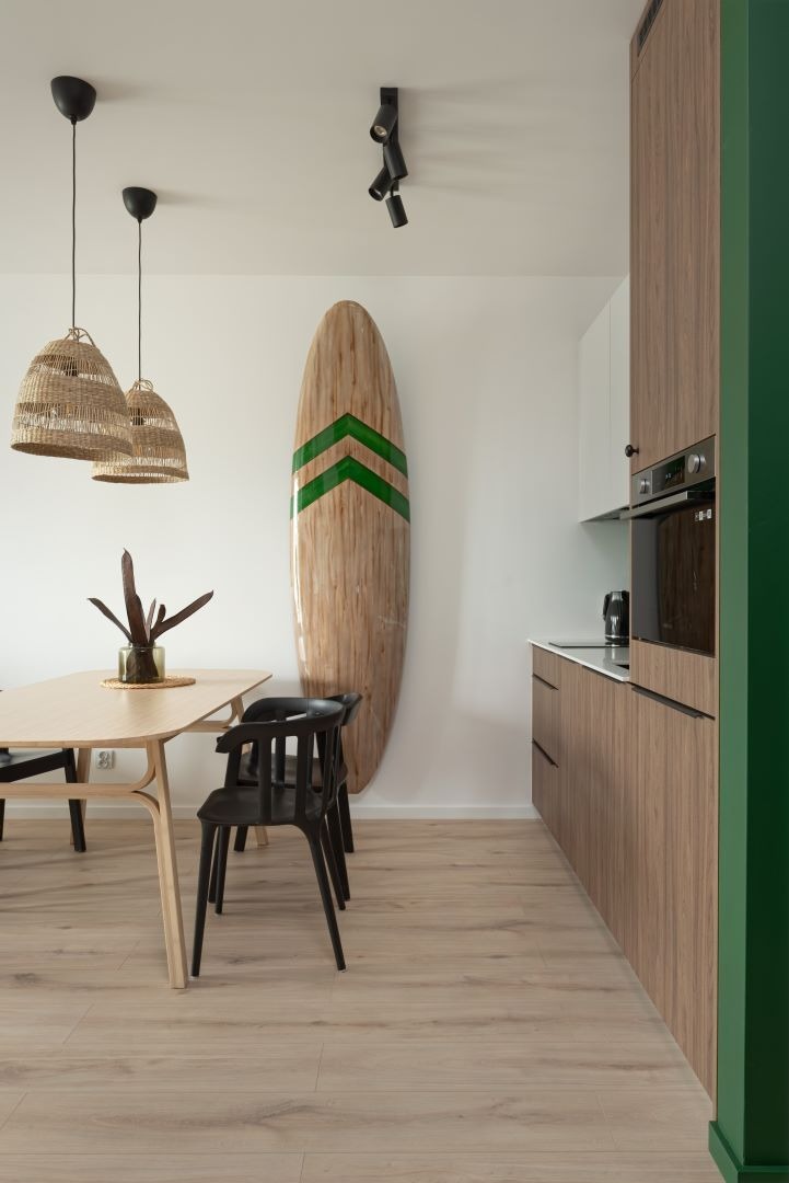 Jasna jadalnia z drewnianym stołem, czarnymi krzesłami, rattanowymi lampami i dekoracyjną deską surfingową z zielonymi pasami opartą o ścianę, sąsiadująca z zabudową kuchenną w odcieniach drewna...