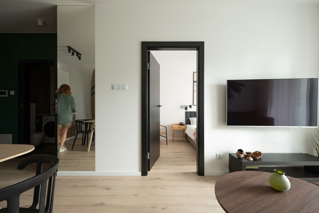 Nowoczesne wnętrze mieszkania z otwartą przestrzenią, widok na sypialnię przez czarne drzwi, telewizor na ścianie, lustro odbijające kuchnię i osobę.