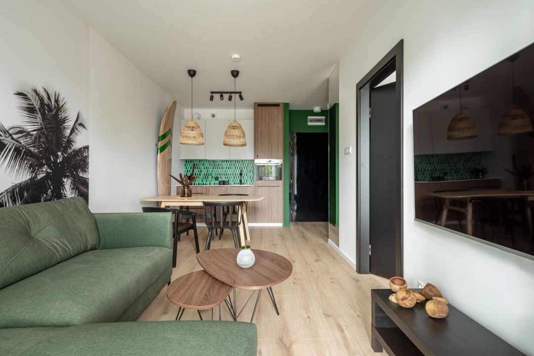 Jasne wnętrze mieszkania w stylu tropikalnym z zieloną sofą, drewnianymi stolikami kawowymi, deską surfingową i plakatem z palmą na ścianie, widok na kuchnię z zielonymi płytkami i drewnianymi...