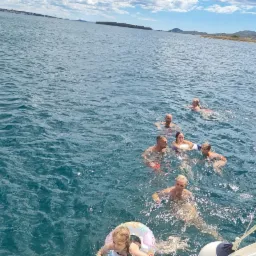 Grupa osób pływa w morzu podczas wycieczki łodzią, z widokiem na oddalony ląd i błękitne niebo z chmurami.