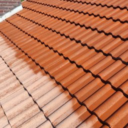 Czyste Dachy - Częściowo odnowiony dach z ceglastą dachówką, widoczny komin i zieleń w tle. Połowa dachu po renowacji, druga połowa w stanie pierwotnym.