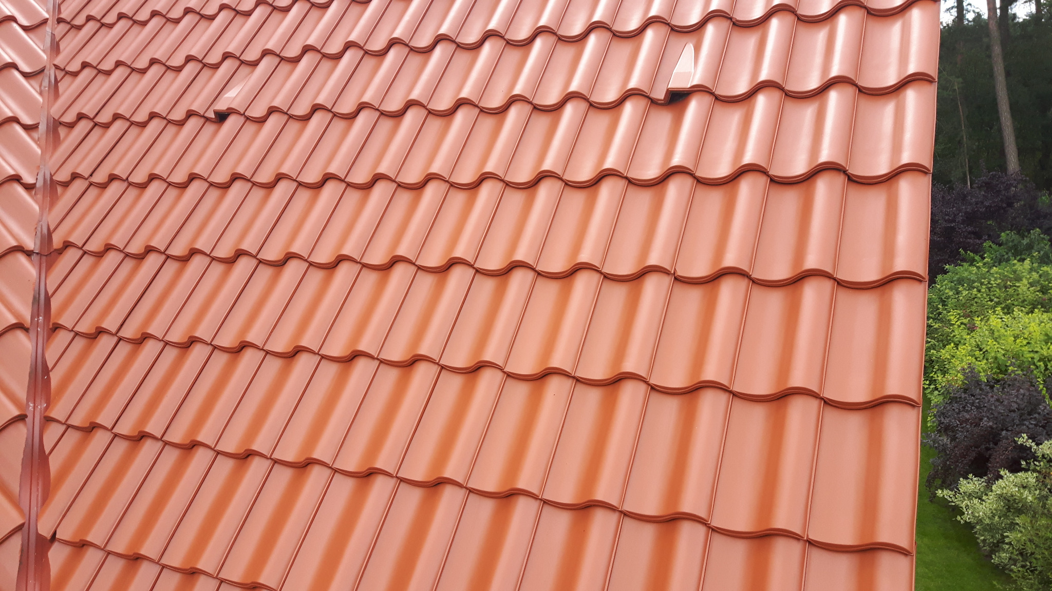 Czerwona dachówka ceramiczna w kształcie fali, widok z góry na fragment dachu z jednym elementem wentylacyjnym, w tle drzewa i krzewy.