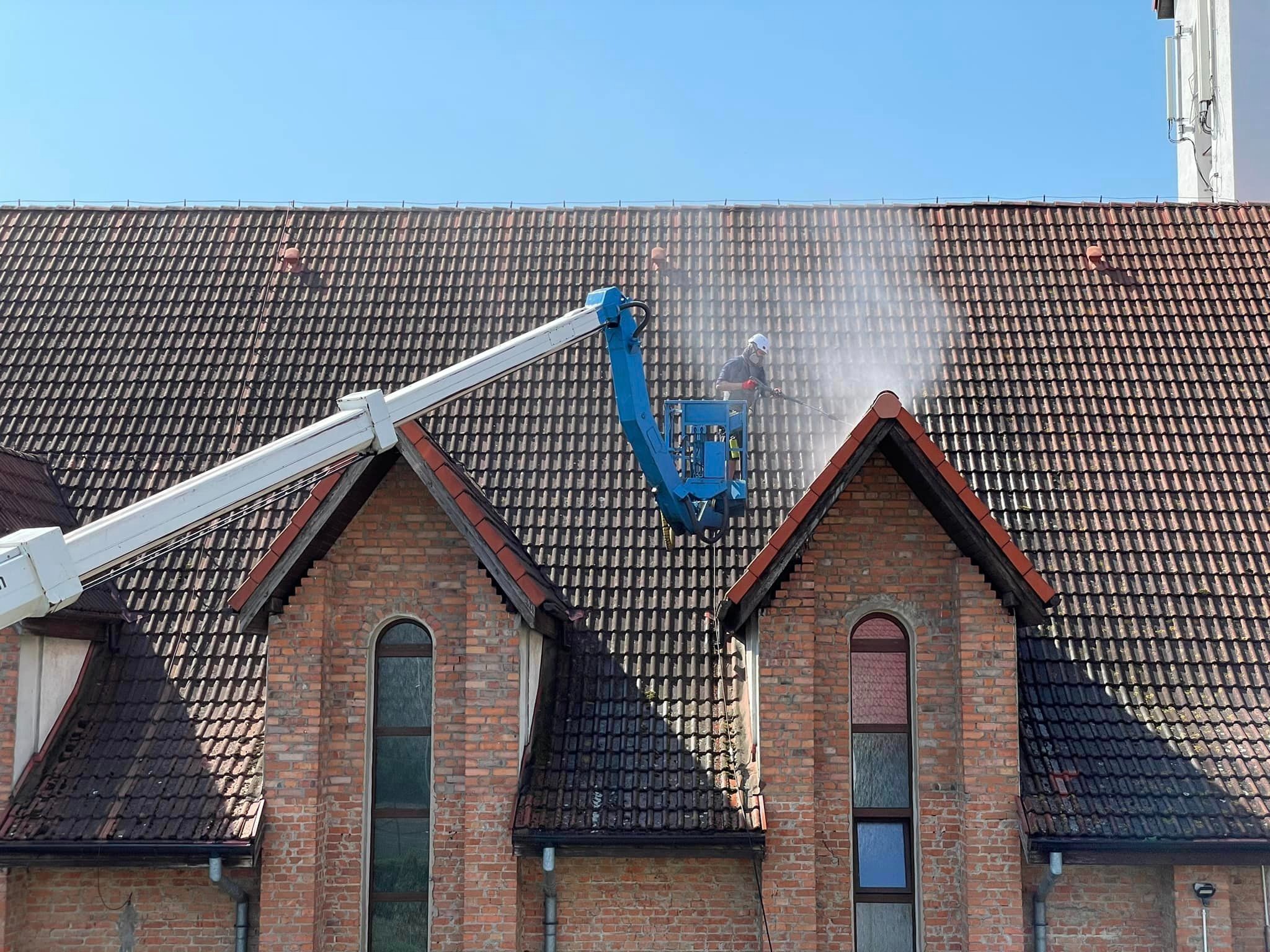 Pracownik na podnośniku czyści dach kościoła z ciemnego nalotu za pomocą myjki ciśnieniowej. Widoczna różnica w czystości połaci dachu. Ceglana fasada budynku.