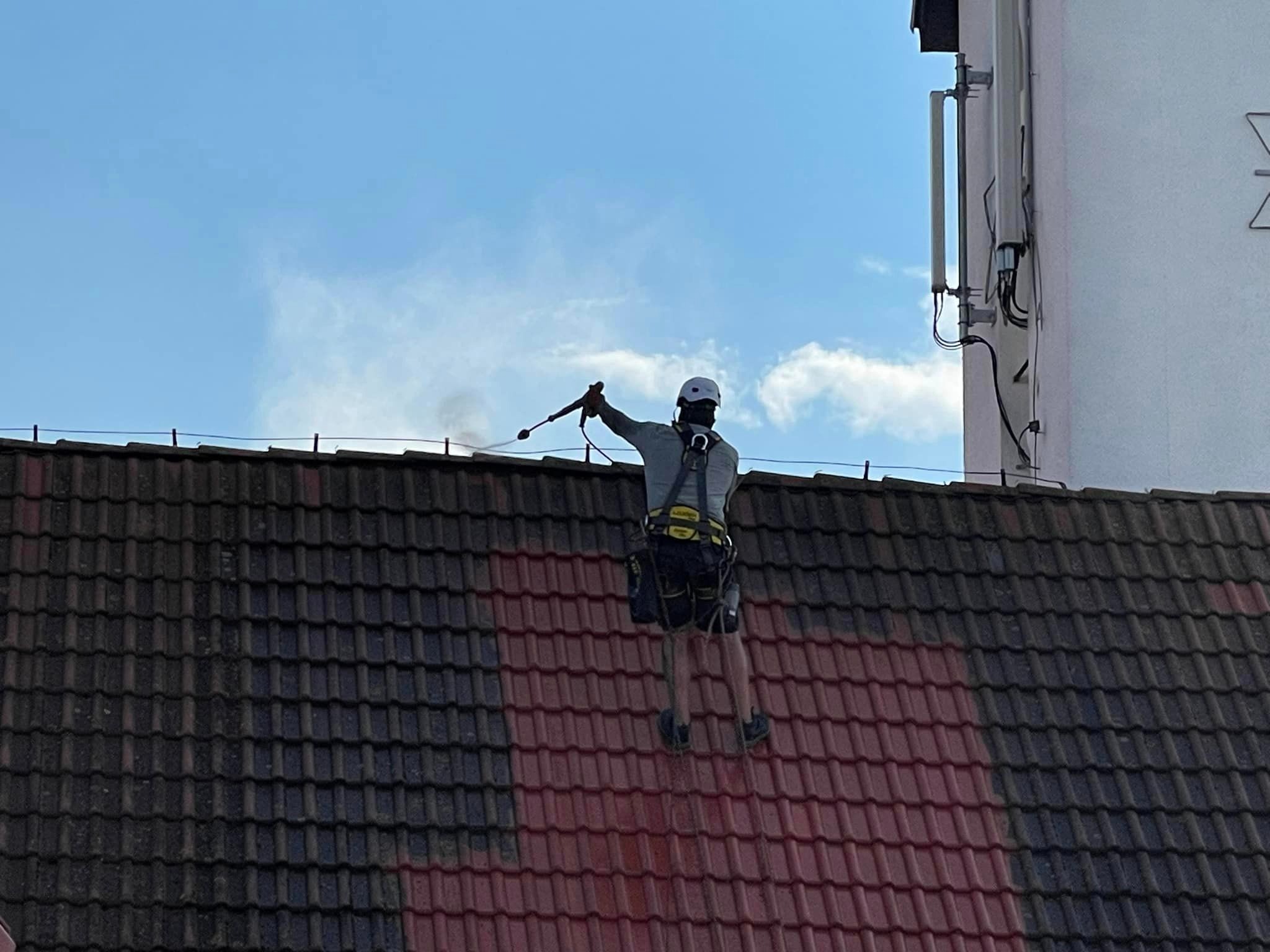 Pracownik w uprzęży, z bliska, czyści dach z ciemnej dachówki za pomocą myjki ciśnieniowej. Część dachu jest już umyta, ukazując czerwoną barwę dachówki.