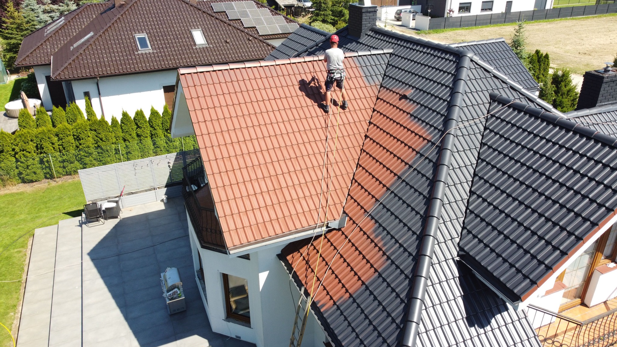 Pracownik na dachu domu w trakcie malowania. Część dachu pomalowana na szaro, część w kolorze ceglastym. Widok z góry, dom z ogrodem w tle.
