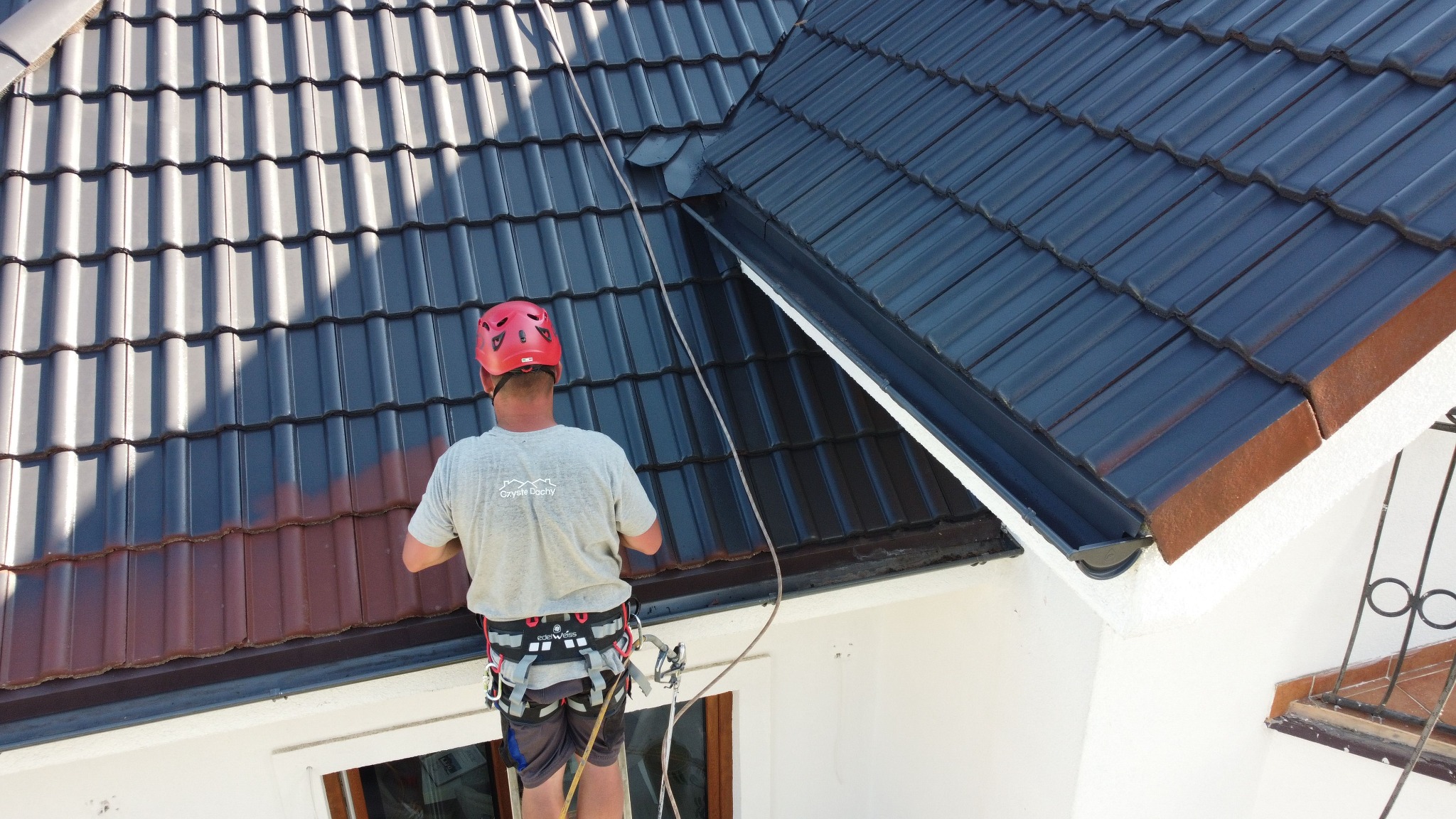 Pracownik w kasku i uprzęży na dachu z dachówki ceramicznej, częściowo umytej, z widoczną różnicą koloru. Ujęcie z góry, w słoneczny dzień.
