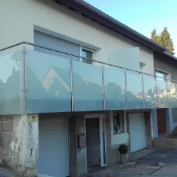 Balkon z przeszkloną balustradą na piętrze domu z garażami w przyziemiu, widok na otoczenie odbity w szkle.