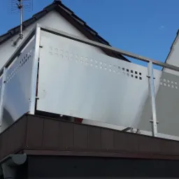 Balkon z balustradą nierdzewną z perforowanymi panelami, widok z dołu na tle niebieskiego nieba i białej ściany budynku