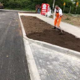 Pracownik w pomarańczowym kombinezonie wyrównuje ziemię grabiami na przygotowanym terenie obok nowej drogi asfaltowej i chodnika z kostki brukowej.