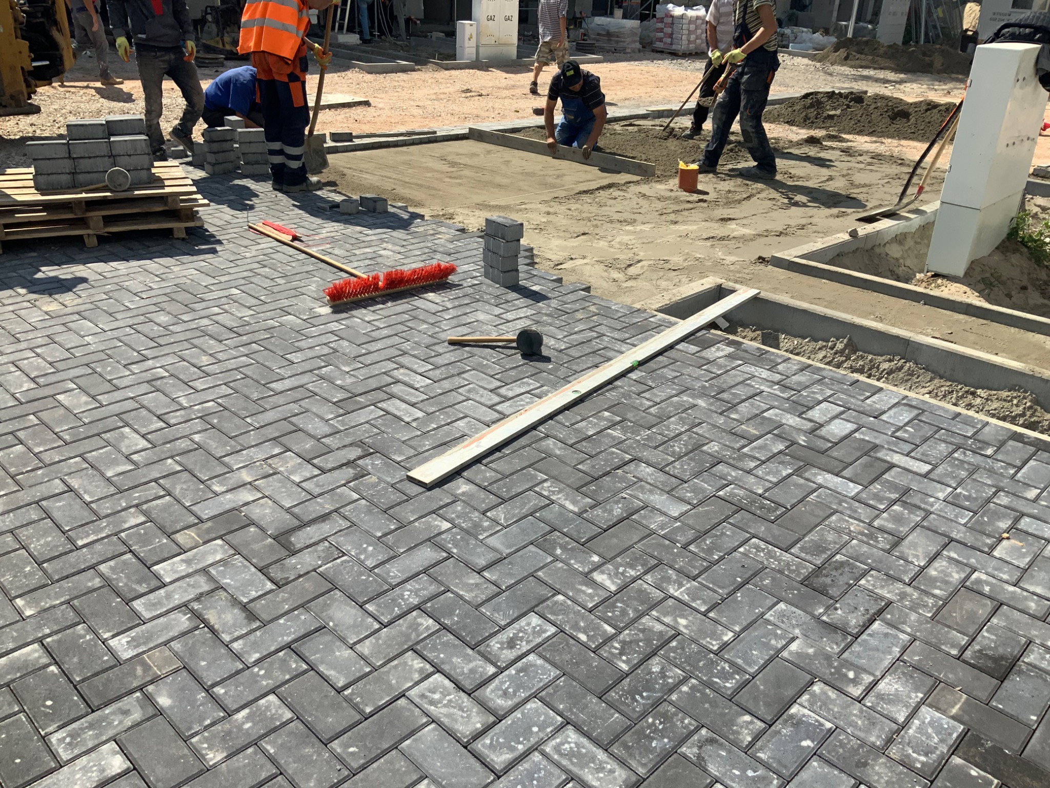 Układanie szarej kostki brukowej w jodełkę, widoczne narzędzia: gumowy młotek i szczotka, w tle pracownicy przygotowujący podłoże.