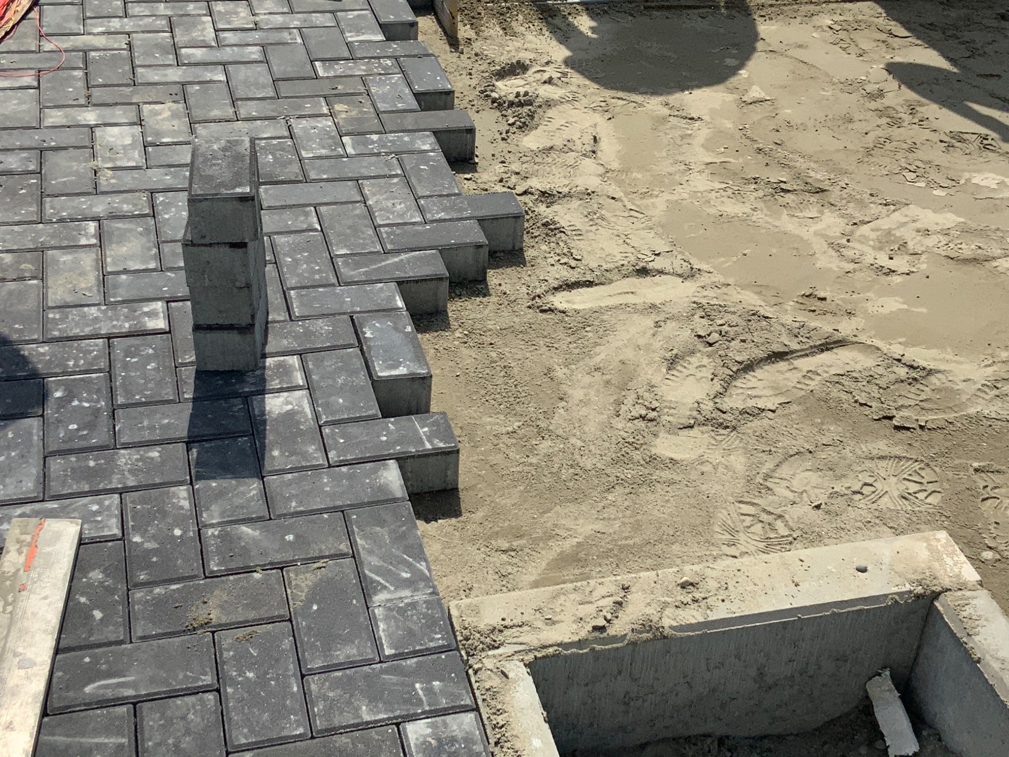 Częściowo ułożona, szara kostka brukowa z widocznym wykopem i betonową studzienką w tle, etap prac brukarskich.