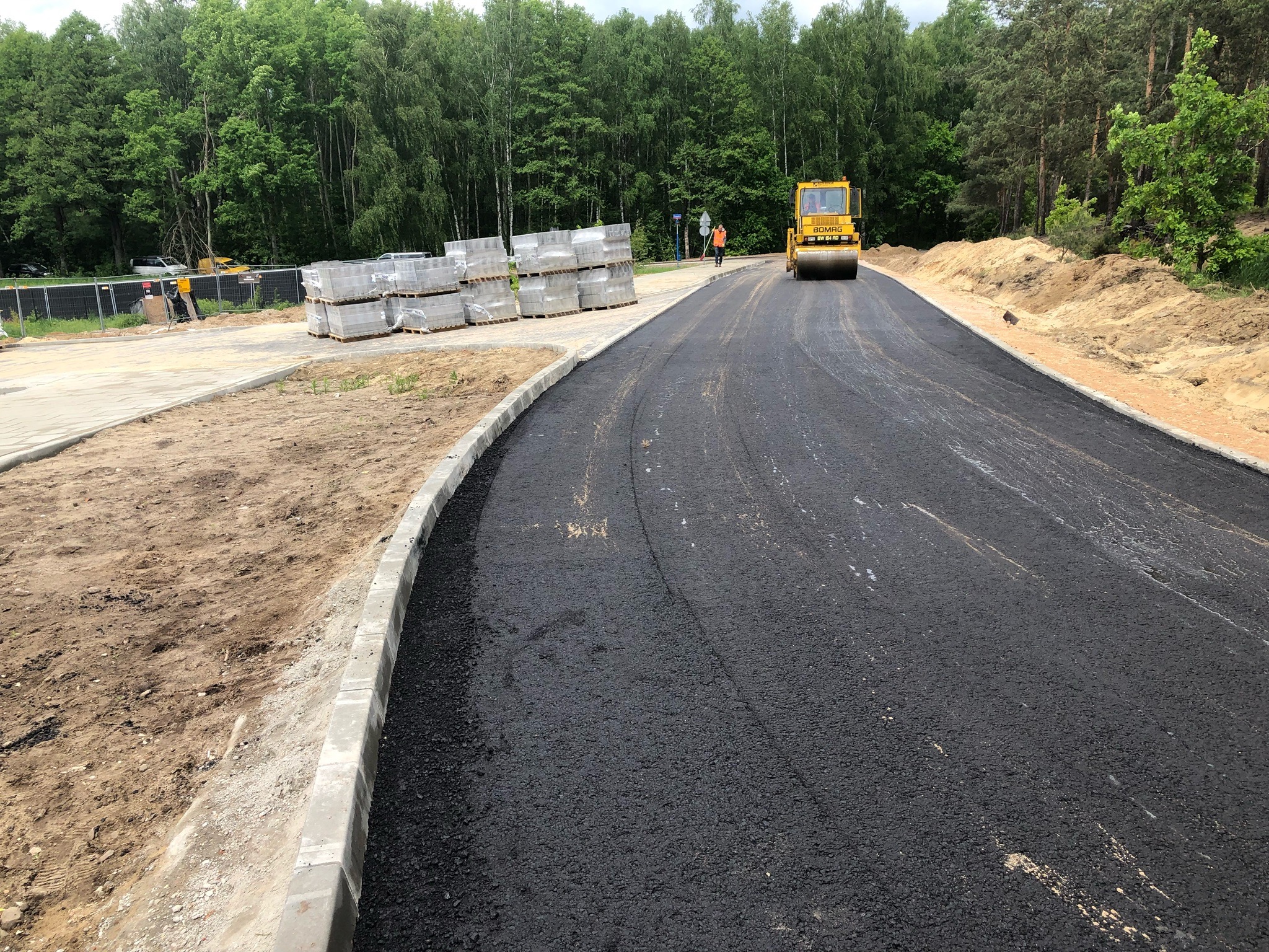 Świeżo ułożona, czarna nawierzchnia asfaltowa drogi w trakcie walcowania przez żółty walec Bomag, z widocznymi paletami kostki brukowej w tle i pracownikiem w pomarańczowej kamizelce.