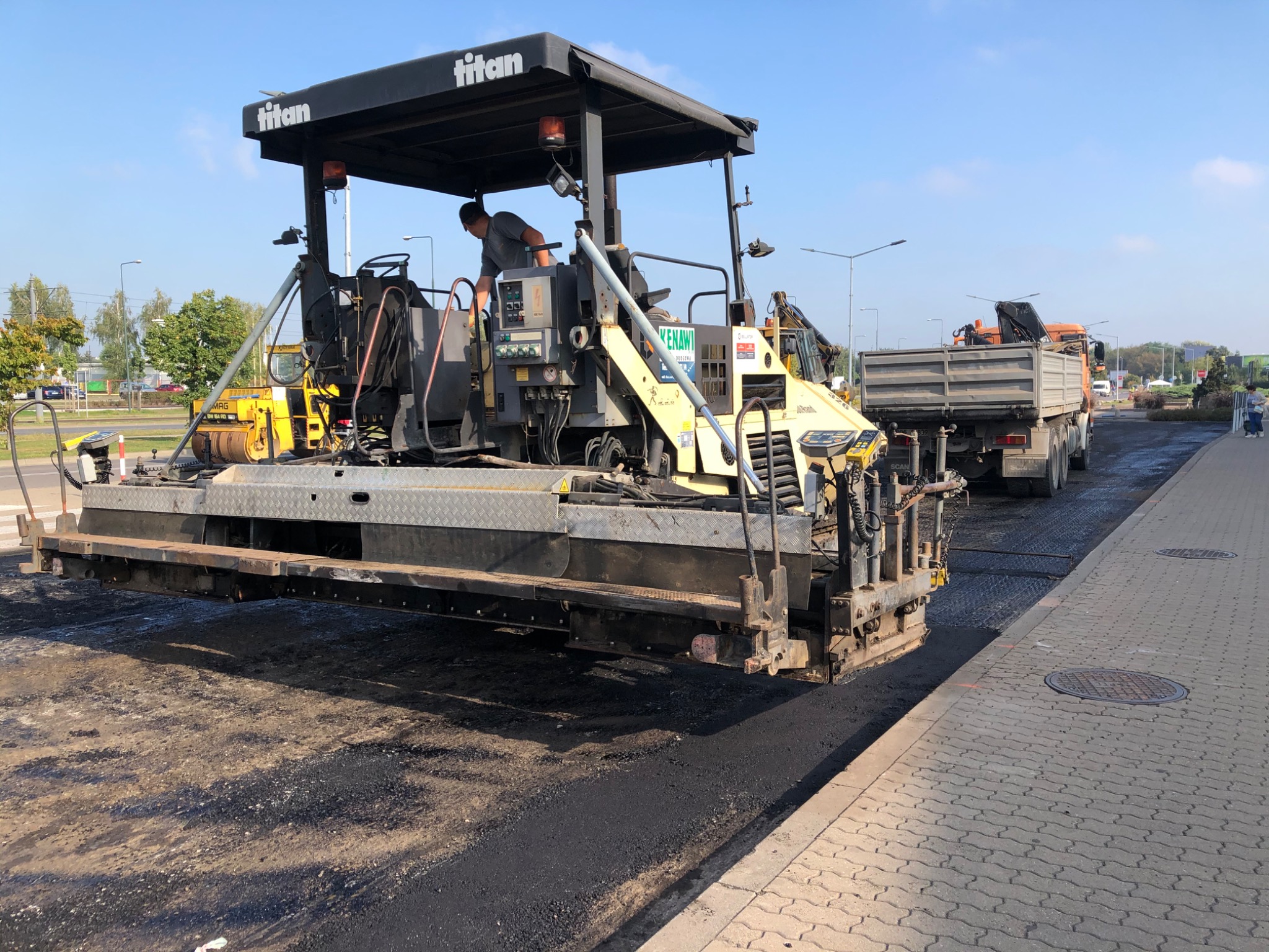 Rozkładanie świeżego asfaltu na drodze przy użyciu specjalistycznej maszyny Titan, widoczny operator i ciężarówka z materiałem w tle.